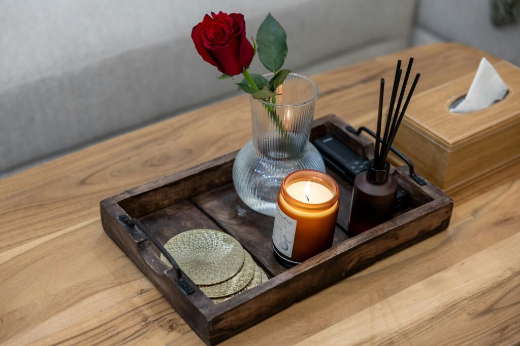 Wooden tray with candle, red rose in vase, reed diffuser, and coasters on a table.